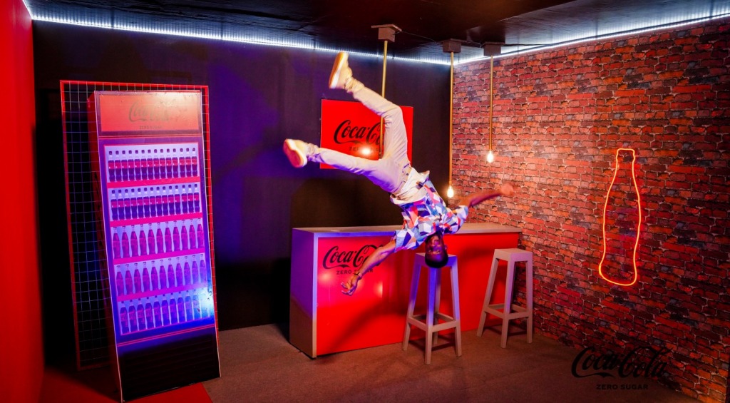 A performer upside down above a Coca-Cola bar, beside a lit fridge and a neon bottle sign