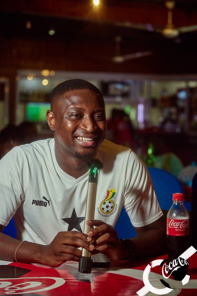 A man in a Ghana football shirt seated at a Coca-Cola branded table