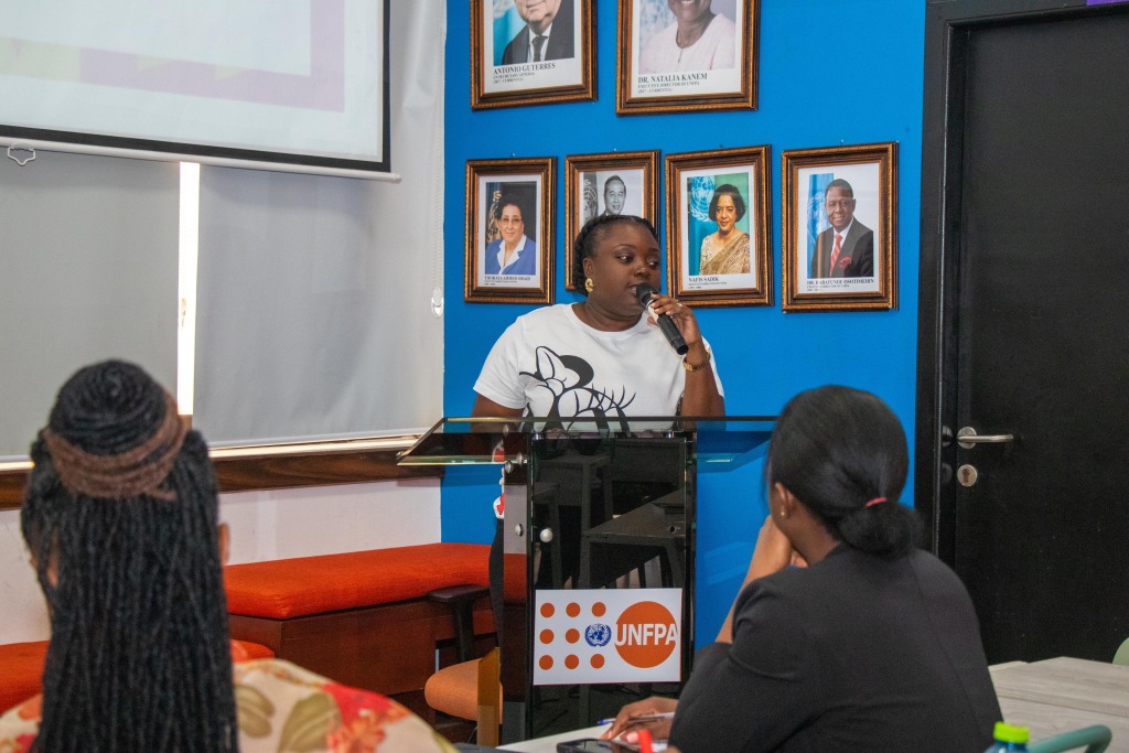 A speaker addressing seated participants at a UNFPA session, framed portraits on the wall behind her